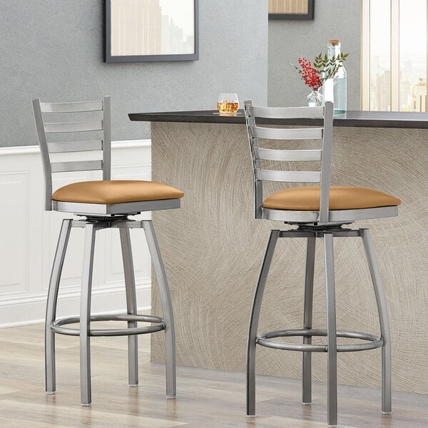 Two Lancaster Table & Seating ladder back swivel bar stools with light brown vinyl padded seats next to a counter.