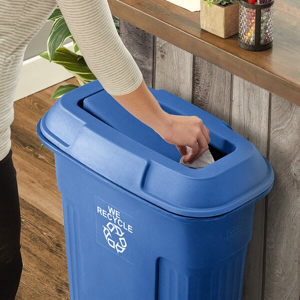 A woman using a blue Toter rectangular lid with swing door to put paper in a trash can.