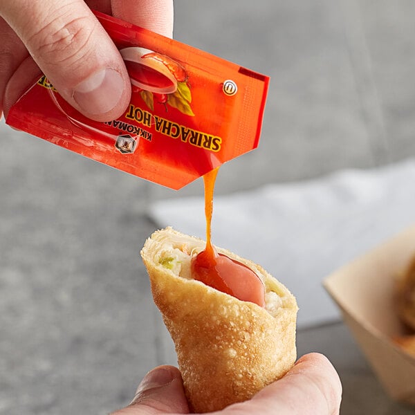 A hand holding a Kikkoman Sriracha hot chili sauce packet over a bowl of sauce.