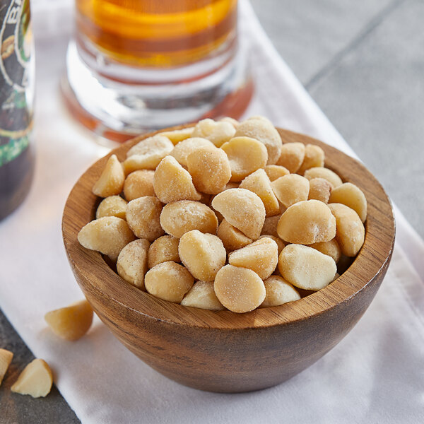 A bowl of dry roasted salted macadamia nuts on a table with a glass of beer.
