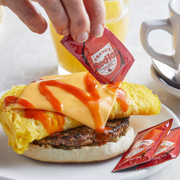 A hand holding a Frank's RedHot Original Cayenne Pepper hot sauce packet over a burger.