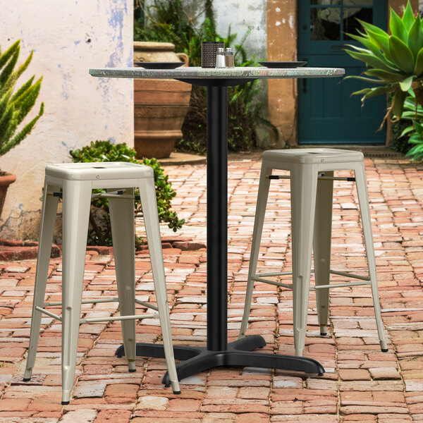A Lancaster Table & Seating black outdoor table base with a table and white stools on a brick patio.