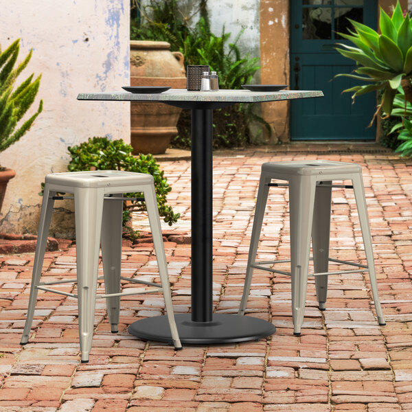 A black Lancaster Table & Seating Excalibur outdoor table base with two white stools on a brick patio.
