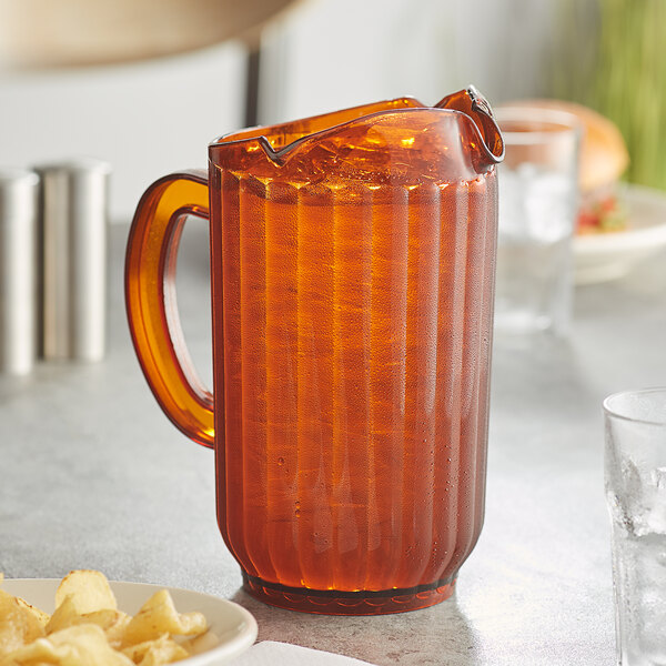 An amber Choice plastic beverage pitcher on a table with a glass of orange juice and a plate of chips.
