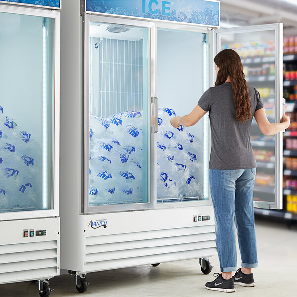 A woman standing in front of a white Avantco glass door ice merchandiser with a large bag of ice.