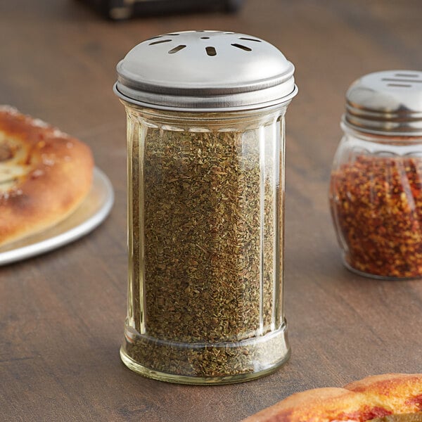 A glass American Metalcraft spice shaker with a stainless steel lid filled with dried herbs on a table alongside a pizza.