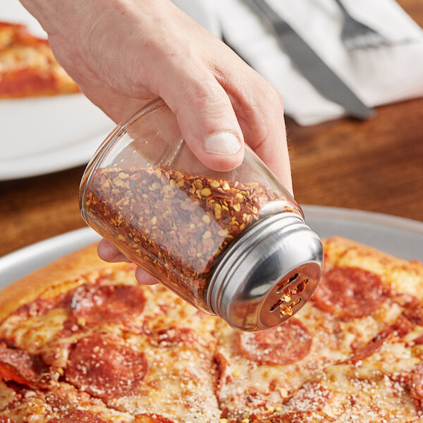 A hand using an American Metalcraft glass spice shaker to sprinkle red pepper flakes on a pizza.