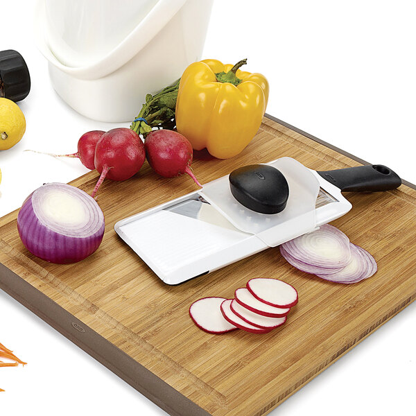 A cutting board with sliced vegetables and an OXO handheld mandoline slicer.