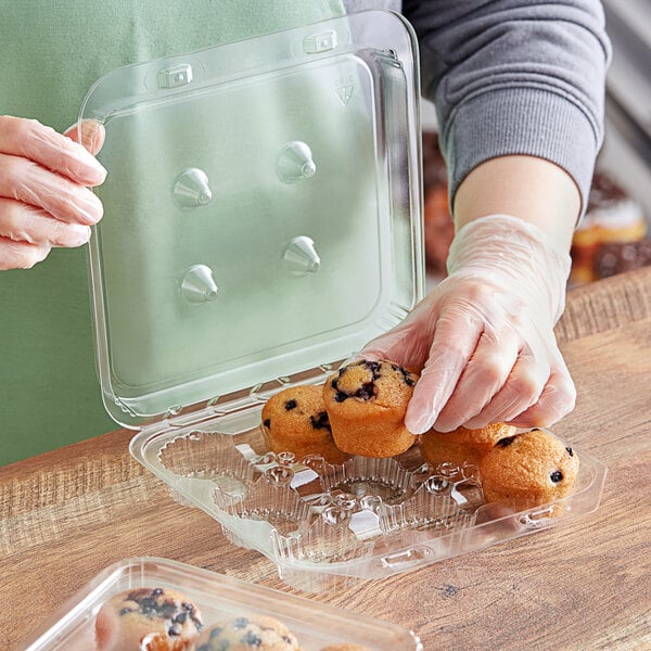 A person in plastic gloves placing a plastic container of muffins on a counter.