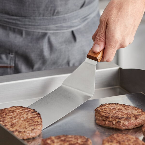 A person using a Vollrath hamburger turner with a wooden handle to flip hamburger patties on a metal surface.