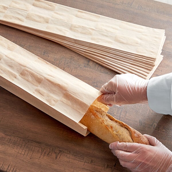 A person wearing gloves putting a baguette in a Bagcraft Packaging paper bread bag.