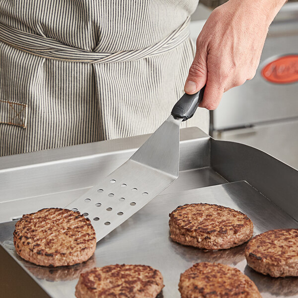 A person using a Vollrath Jacob's Pride perforated turner to cook burgers on a grill.