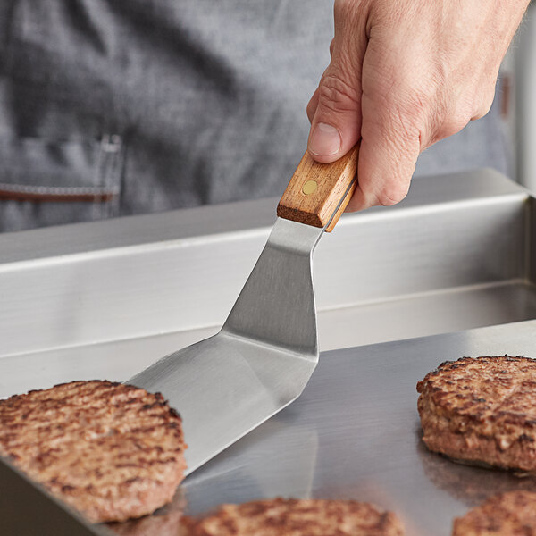 A person using a Vollrath stainless steel hamburger turner with a wooden handle to cook hamburger patties.