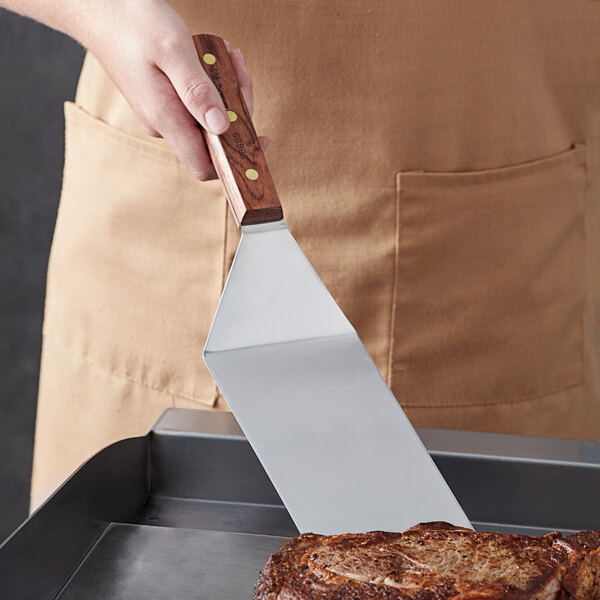 A person holding a Dexter-Russell solid steak turner over a steak on a grill.