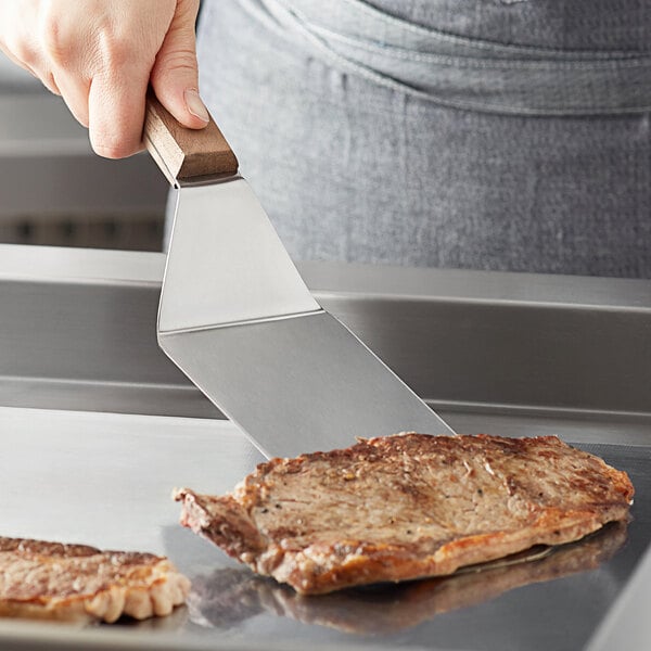 A person using a Dexter-Russell solid steak turner to flip a piece of meat on a grill.