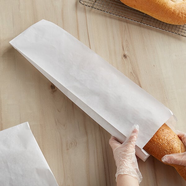 A gloved hand holds a Bagcraft white paper bag with a loaf of bread inside.