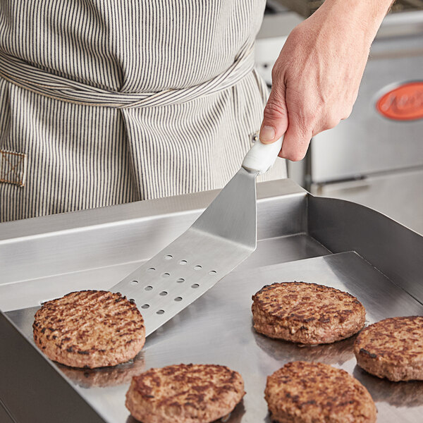 A person using a Vollrath Jacob's Pride perforated high heat turner to cook hamburgers on a grill.