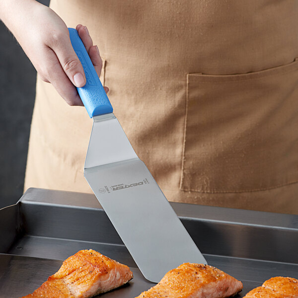 A hand holding a Dexter-Russell solid turner with a blue plastic handle over a piece of cooked salmon.