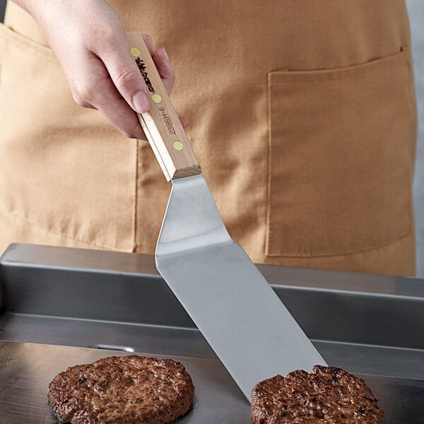 A hand holding a Dexter-Russell high-carbon steel turner with a wooden handle cooking burgers on a grill.