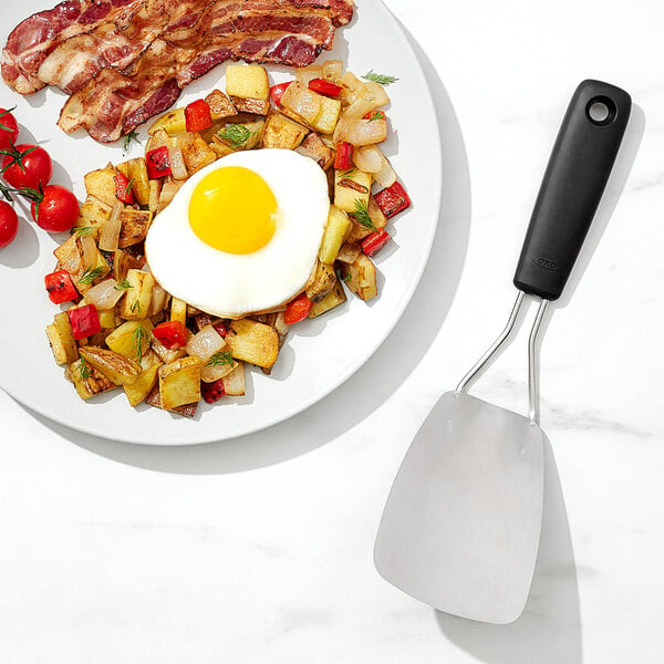 A plate of food with eggs, bacon, and potatoes next to an OXO stainless steel spatula.