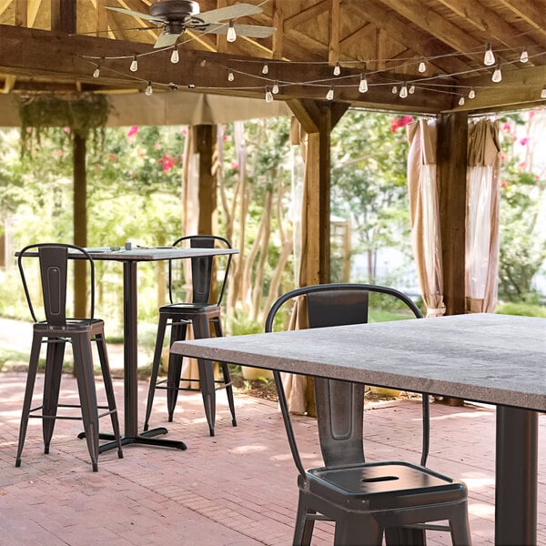 A Lancaster Table & Seating Excalibur bar height table with a textured Toscano finish and cross base plate on a covered patio with chairs.