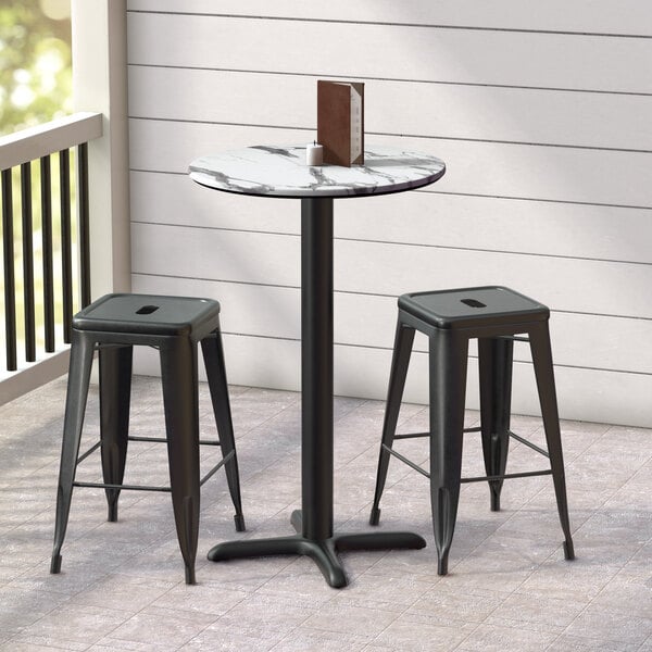 A Lancaster Table & Seating round counter height table with cross base plate and two black bar stools on a porch.
