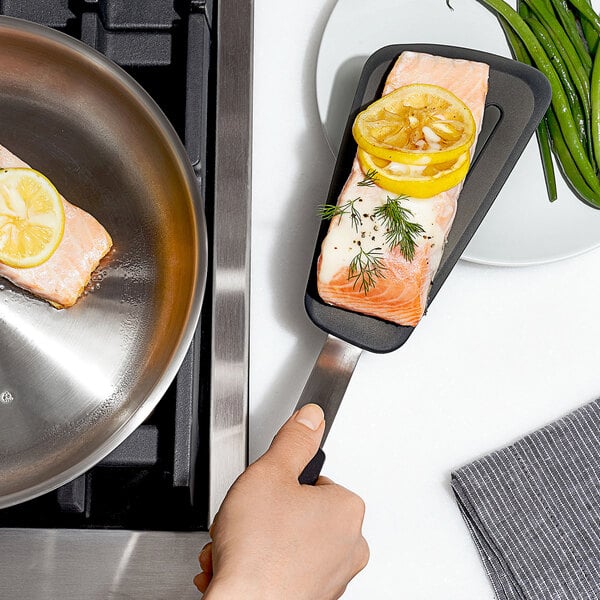 A hand holding a black OXO silicone slotted spatula over a pan with salmon.