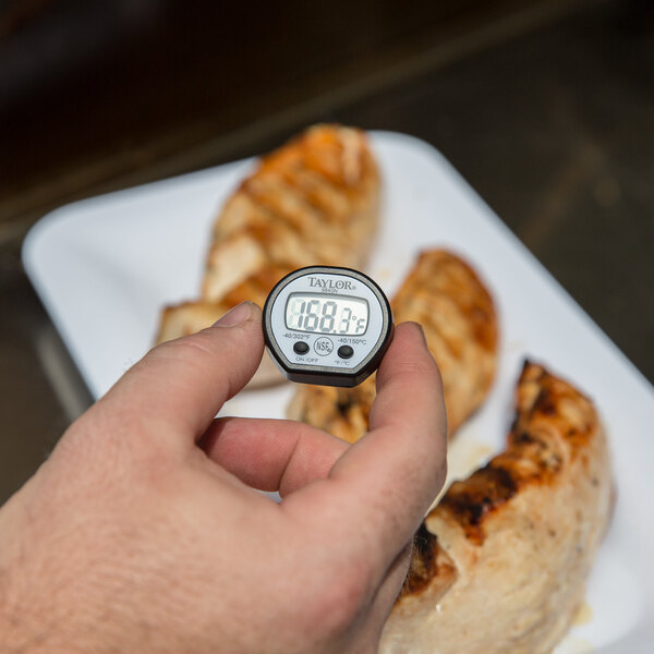 A hand holding a Taylor digital pocket probe thermometer over a plate of food.