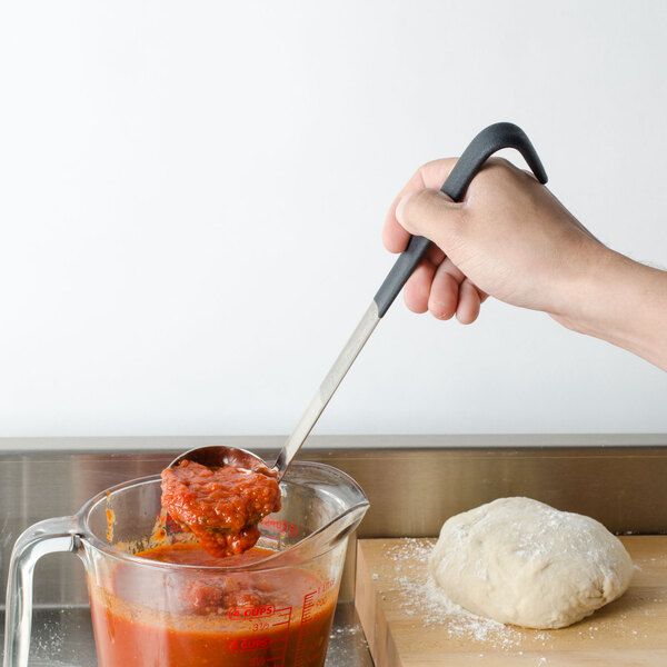 A person's hand holding a Vollrath stainless steel ladle with a black handle over a bowl of sauce.