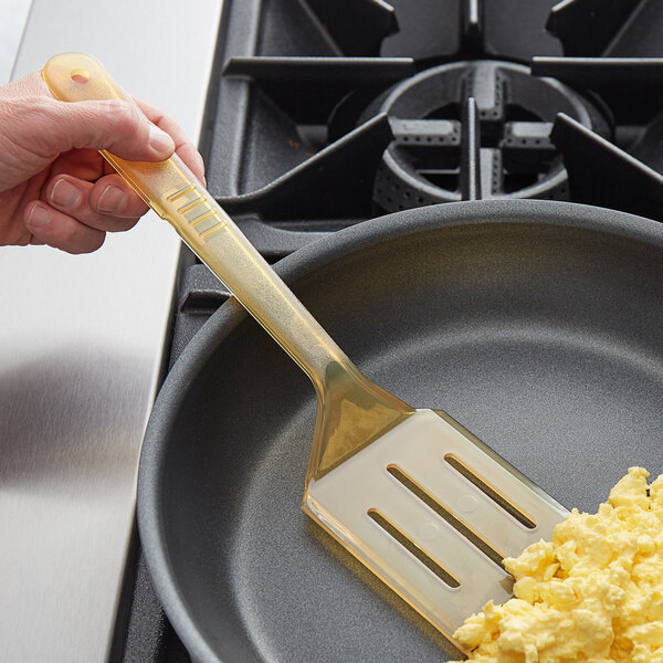 A hand holding a yellow plastic Carlisle slotted spatula over scrambled eggs in a frying pan.