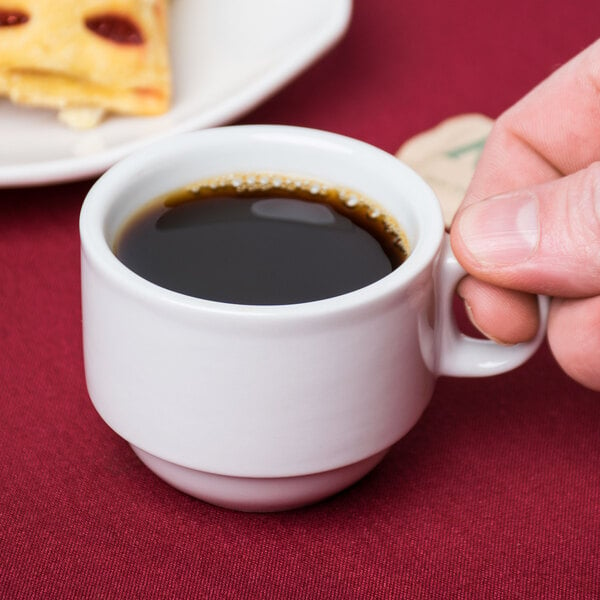 A hand holding a Tuxton bright white espresso cup filled with coffee on a table.