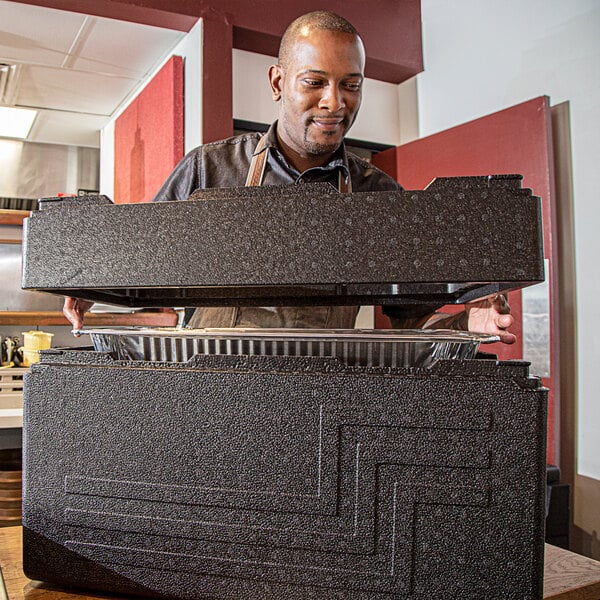 A man standing in a kitchen with a Cambro Cam GoBox top-loading black food pan carrier.