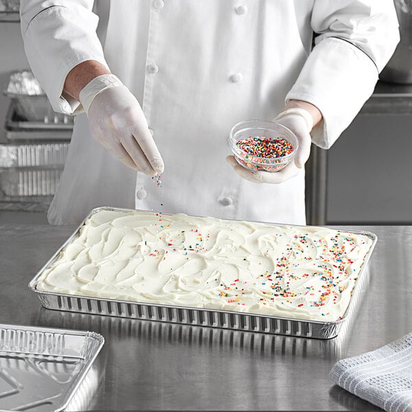 A person in a white coat and gloves sprinkles sprinkles on a frosted cake in a Baker's Mark foil cake pan.