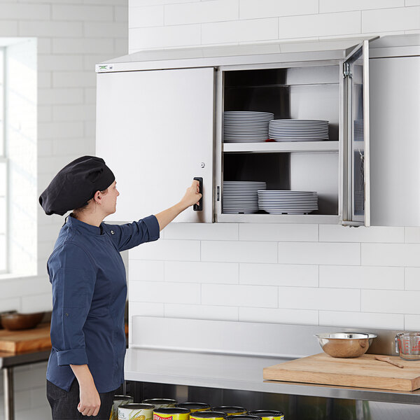 A woman in a chef hat opening a Regency stainless steel wall cabinet with plates in it.