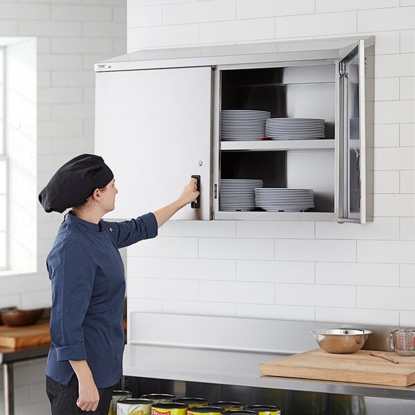A woman wearing a chef hat opening a Regency stainless steel wall cabinet with plates in it.