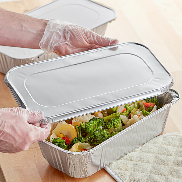 A person wearing a plastic glove places a Choice third size foil steam table pan lid on a tray of food.