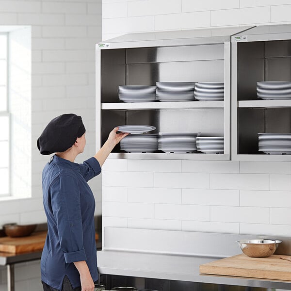 A woman in a chef's hat puts plates on a Regency stainless steel wall cabinet.