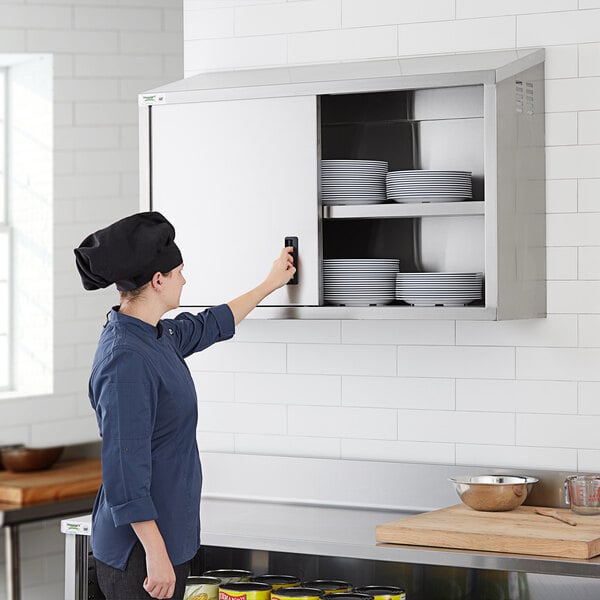 A woman in a professional kitchen opening a Regency stainless steel wall cabinet with sliding doors to access plates.