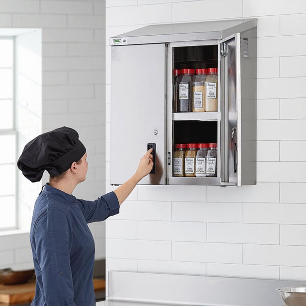 A woman in a chef hat opening a Regency stainless steel wall cabinet with hinged doors to access food items.