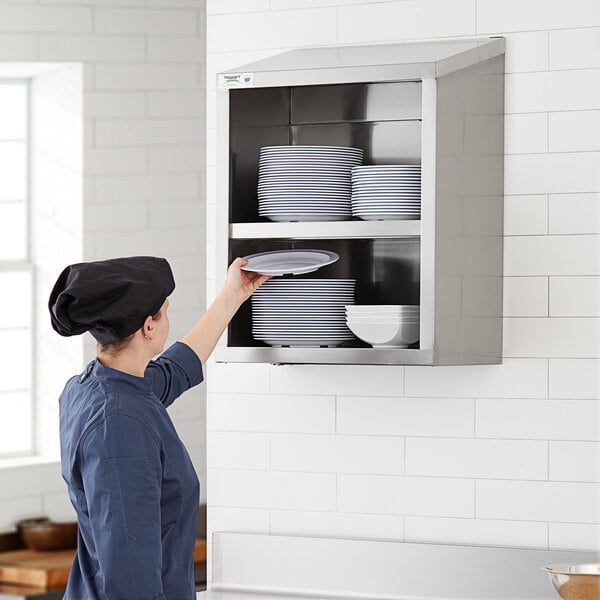 A woman wearing a chef hat putting plates into a Regency stainless steel wall cabinet.