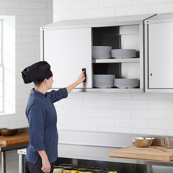 A woman in a professional kitchen opening a Regency stainless steel wall cabinet with sliding doors.