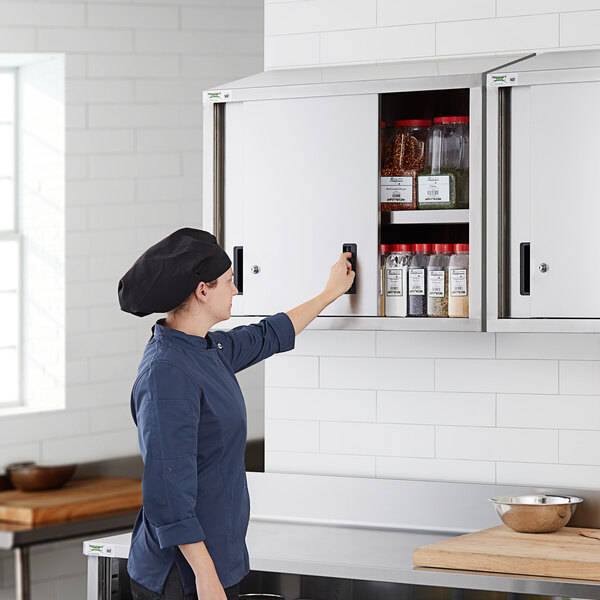 A woman wearing a chef hat opening a Regency stainless steel wall cabinet filled with spices.
