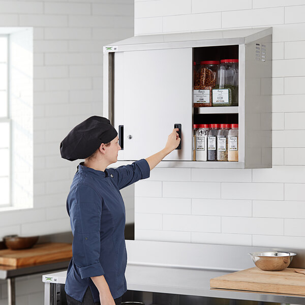A woman in a chef hat opening a Regency stainless steel wall cabinet in a professional kitchen.