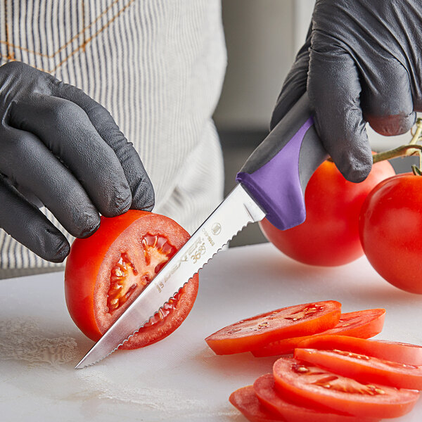 A person in black gloves using a Dexter-Russell purple scalloped utility knife to cut a tomato.