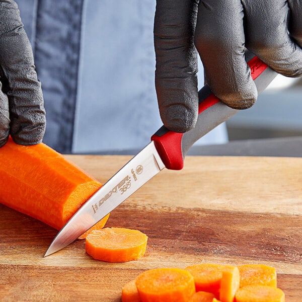 A hand holding a Dexter-Russell paring knife cutting a carrot on a wooden cutting board.