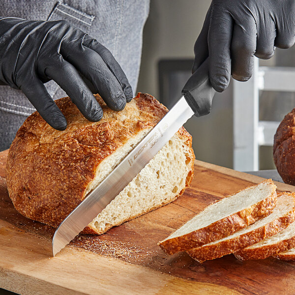 A person wearing black gloves uses a Dexter-Russell scalloped bread knife to slice bread on a cutting board.
