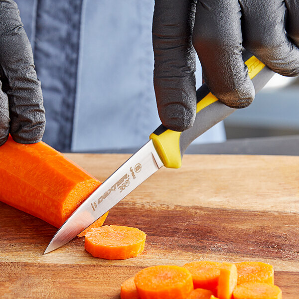 A person in black gloves using a Dexter-Russell paring knife with a yellow and black handle to cut a carrot on a wooden cutting board.