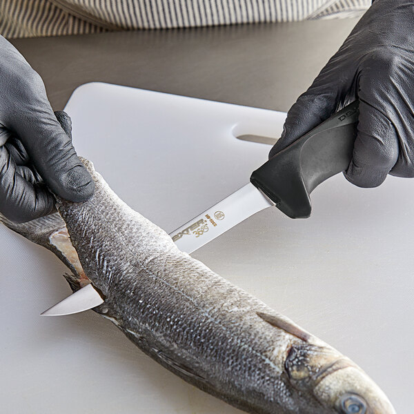A person in a black glove using a Dexter-Russell narrow boning knife to cut a fish on a cutting board.