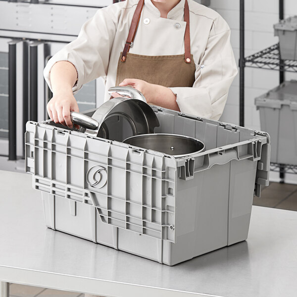 A woman in a chef's uniform holding a gray Orbis Stack-N-Nest Flipak tote box.