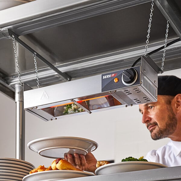 A man holding a plate of food under a ServIt strip warmer in a professional kitchen.